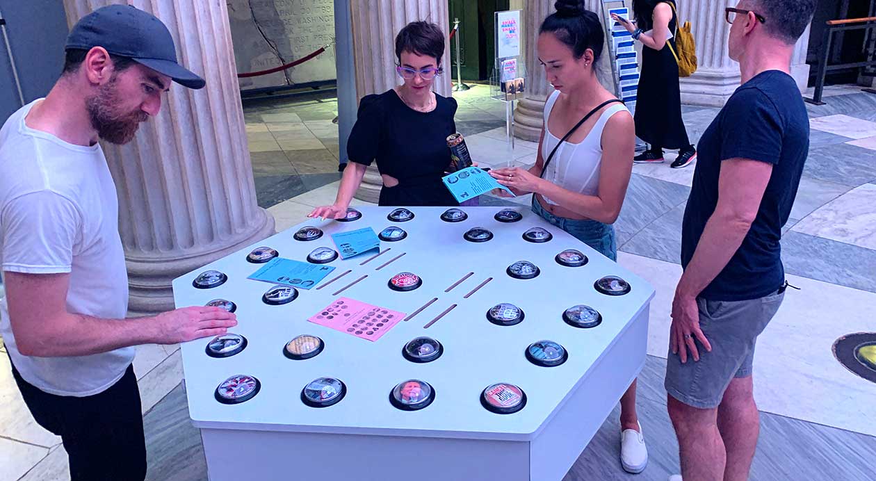 Four people gathered around the work, with two of the players touching one of the buttons