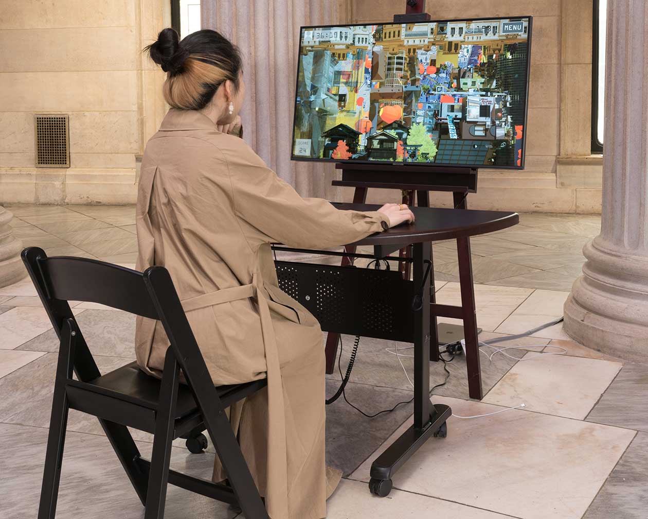 Person seated at desk with keyboard and mouse in front of screen on easel, with the game on the screen