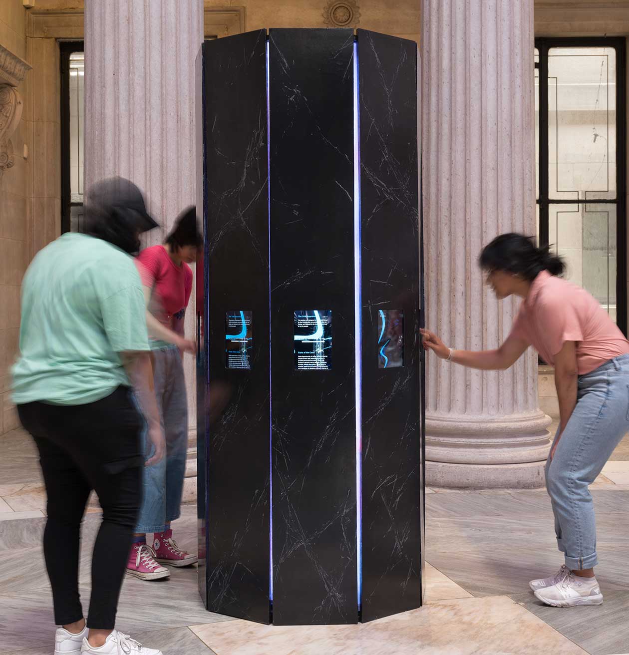 Three people interact with the artwork, v.erses. The work is composed of a 9-sided column in which tablet screens are embedded